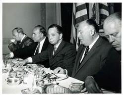["Black and white photograph print of Carl Albert speaking to men seated at a dinner table. Gerald R. Ford is present."]
