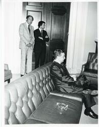 ["Black and white photograph print of Carl Albert seated on a couch. Two unidentified men are standing in the background"]