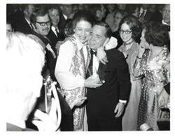 ["Black and white photograph of Carl Albert, Mary Albert, and their daughter Mary Frances are shown in crowd. Carl Albert is being hugged by an unidentified woman."]